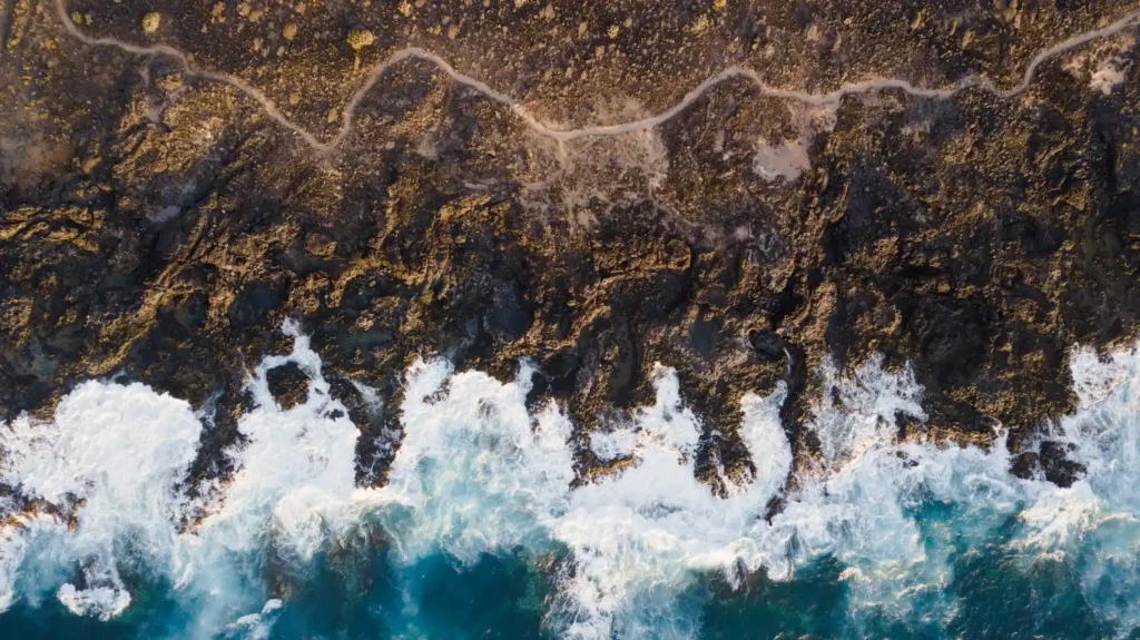 Les bienfaits du magnétisme sur la santé et le corps 6 Vue aérienne des vagues turbulentes s’écrasant sur une côte rocheuse, eau turquoise et mousse blanche. Bienfaits du magnétisme sur la santé évoqués.