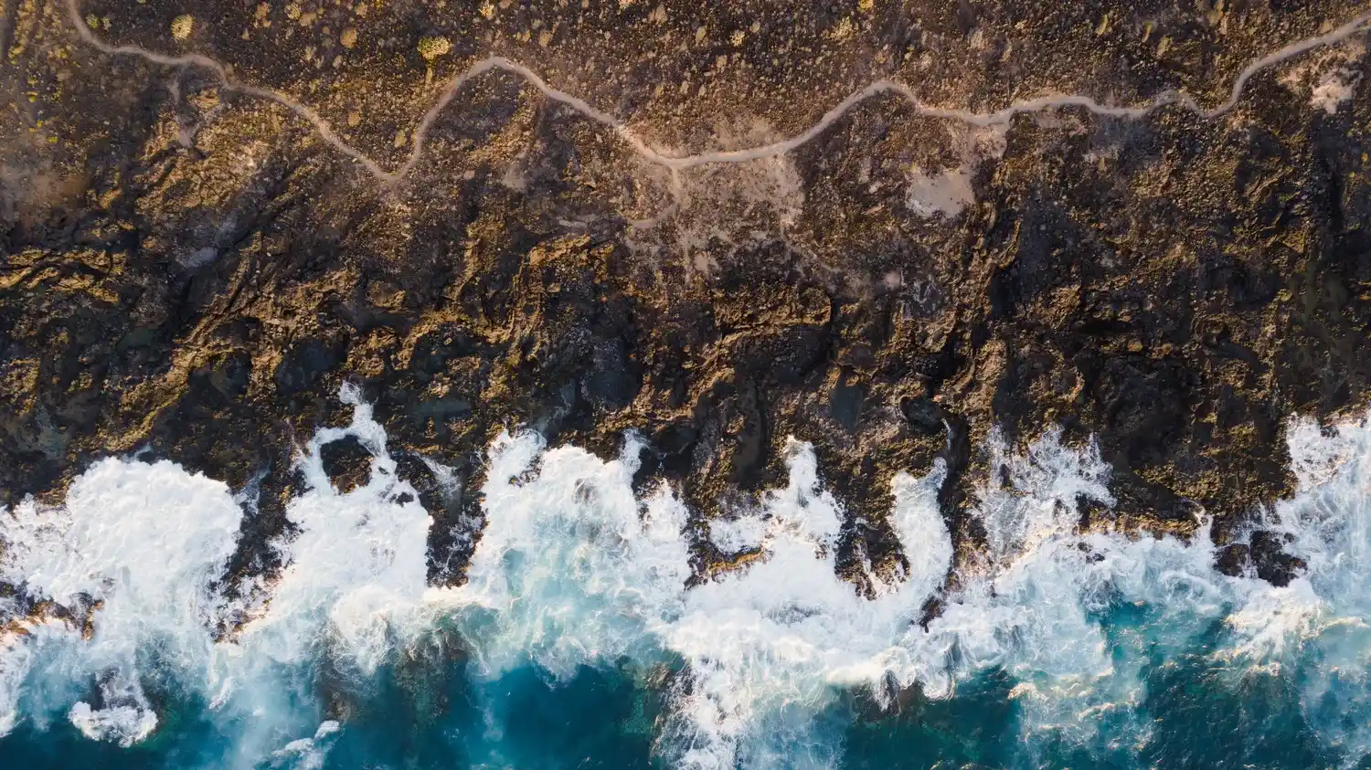 Vue aérienne des vagues turbulentes s’écrasant sur une côte rocheuse, eau turquoise et mousse blanche. Bienfaits du magnétisme sur la santé évoqués.